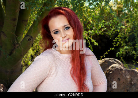 A beautiful, teen-aged girl with dyed red hair poses for a portrait in front of trees.  She has a closed mouth smile. Stock Photo