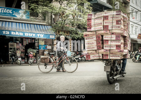 Vietnamese person pushing a traditional bike alongside a heavily ladened motorcyle in a typical street with traffic in Hanoi, Vietnam Stock Photo