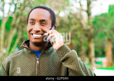 Handsome hispanic black man wearing green sweater in outdoors park area holding up phone to ear and talking happily while laughing Stock Photo