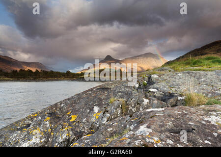 Rainbow arching over the Pap of Glencoe or Sgorr na Ciche during unsettled weather, with granite rocks and Loch Leven at front Stock Photo