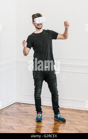 A young man wearing black in the corner of a room using his hands in an animated manner while wearing a Virtual Reality Headset Stock Photo