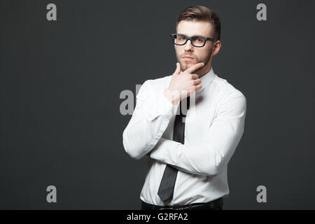Young thoughtful handsome businessman standing against dark gray background. Stock Photo
