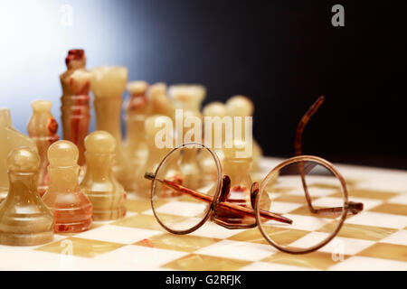 Set of chess pieces made from Onyx near old spectacles on board against dark background Stock Photo