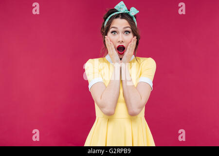 Amazed pretty pinup girl in yellow dress standing with mouth opened over pink background Stock Photo