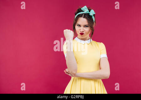 Angry pretty pinup girl in yellow dress showing her fist over pink background Stock Photo