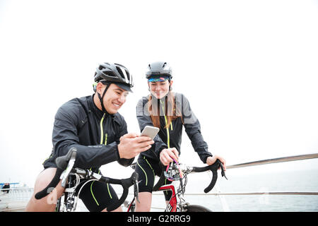 Smiling couple on bicycles resting and using smartphone together outdoors Stock Photo