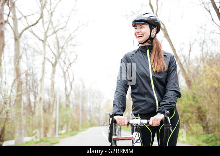 Cheerful beautiful young woman in protective helmet with bike walking in park Stock Photo