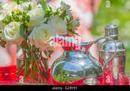 still life with silver pitcher and silver cocktail shaker, fresh cut roses in glass vase with water on red table Stock Photo