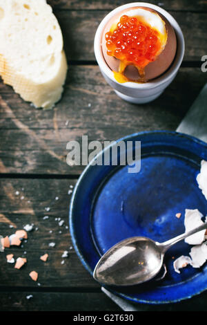 Breakfast with soft-boiled egg with red caviar in white eggcup, served with bread over old wooden table. Focus on caviar Stock Photo