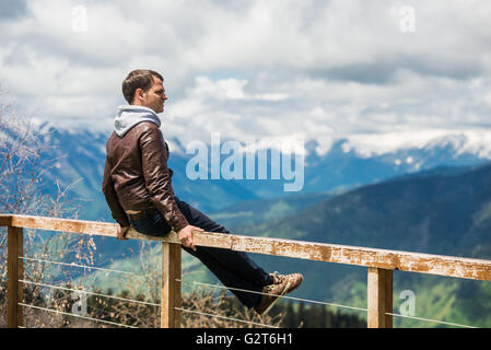 Back view of a man is thinking about this amazing world, while sitting at mountains against jungle view. Young male traveler enjoying beautiful landscape during road trip Stock Photo