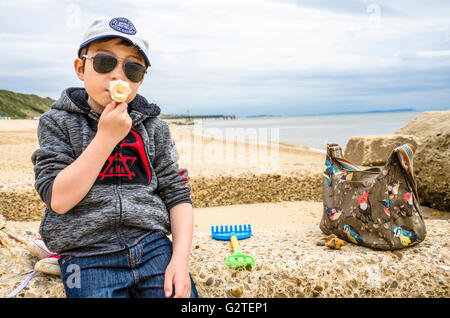 A young boy sits on part of an old sea defense on Boscombe Beach, Bournemouth eating an ice cream. Stock Photo
