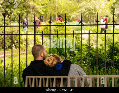 A couple on a bench hugging in front of a public garden and a railing Stock Photo