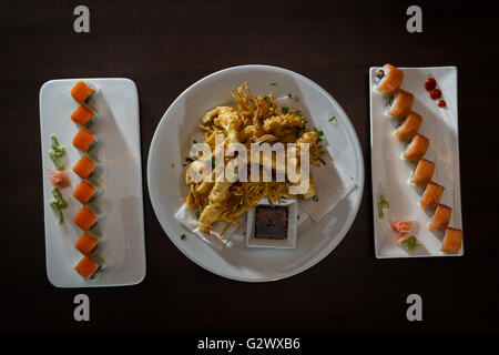 close up of a tempura shrimp surrounded by two fresh sushi rolls served on white plates on a wooden table Stock Photo