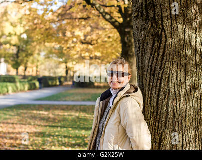 Mature, smiling woman in autumn park at tree, horizontal Stock Photo