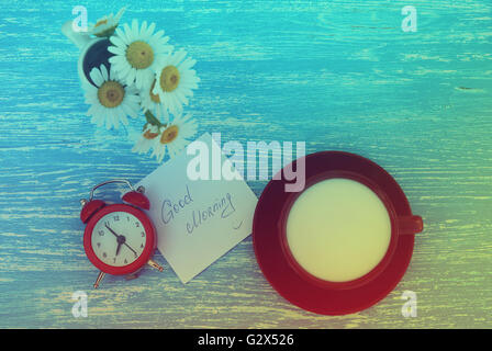 Daisy flowers, alarm clock and cup of milk with good morning note on rustic blue wooden background. Photo in vintage style Stock Photo