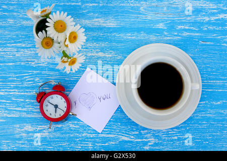 Daisy flowers, alarm clock and cup of coffee with I love you note on rustic blue wooden background Stock Photo