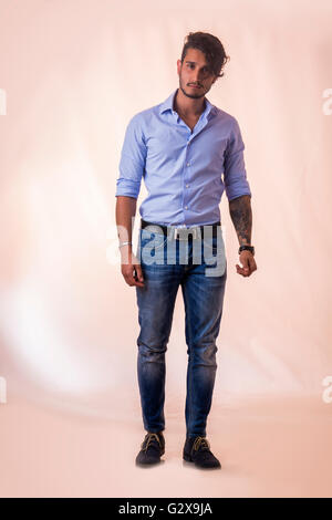 Portrait of brunette young man in light blue shirt and jeans, standing in studio shot against light background. Full length phot Stock Photo