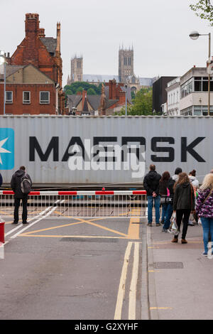 People / pedestrians wait at a railway level crossing barrier in Lincoln, Lincolnshire, UK, as a freight train passes through. Stock Photo