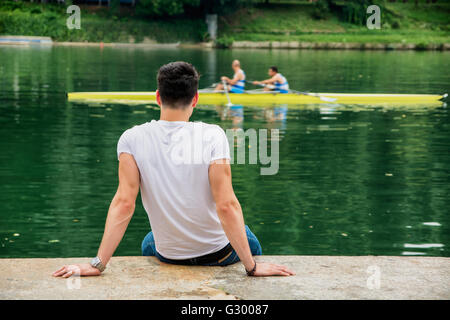 Contemplative light brown haired handsome young man wearing white t-shirt and denim jeans sitting on wall beside picturesque riv Stock Photo