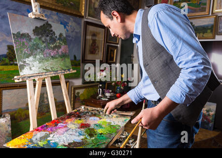 Professional artist working in a gallery or studio selecting a particular hue from a colorful palette of blended oil paints as he works on a painting on an easel. Stock Photo