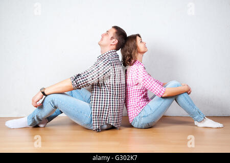 Young couple relaxing on the floor at home sitting barefoot back to back each looking up into the air with a thoughtful smile as Stock Photo