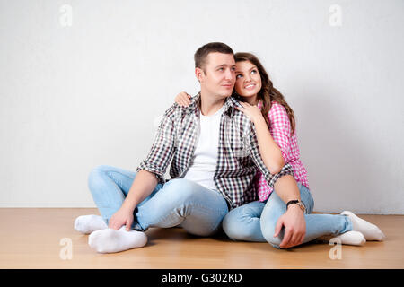 Beautiful barefoot young couple in casual jeans sitting leaning against a white wall on the wooden floor in the living room smil Stock Photo