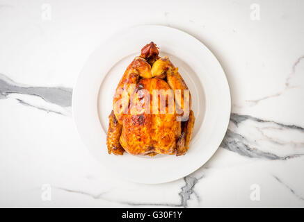 Rotisserie Chicken. Whole roast chicken on white plate seen from above on marble work surface Stock Photo