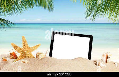 Summer concept with empty tablet display for copyspace. Various kind of shells in sand. Palm leaves on foreground Stock Photo