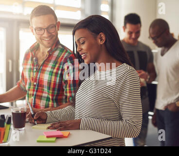 Attractive male wearing eyeglasses and flannel shirt standing next to female co-worker writing on sticky notes at desk with othe Stock Photo