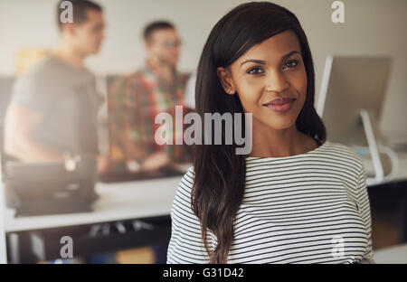 Single beautiful female business owner wearing black and white striped blouse with two employees on computer in background Stock Photo