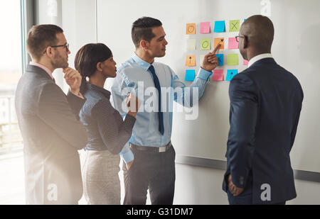 Multiracial group of colleagues discussing a business plan standing around a set of colorful memo notes stuck on the wall Stock Photo