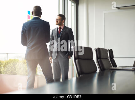 Two multiracial businessmen standing talking together in front of a window in a conference room, view over the table with sun fl Stock Photo