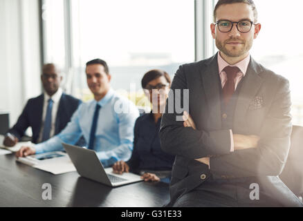 Handsome bearded professional sitting on desk corner beside three coworkers in front of large office window Stock Photo