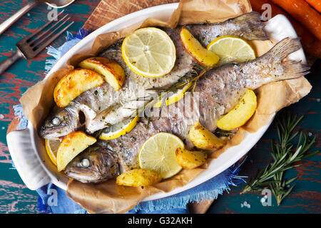 An oven roasted trout in a casserole on a rustic table. With carrots and potatoes on the side. Stock Photo