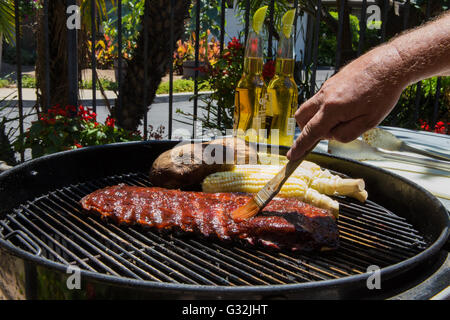 Barbecued baby-back pork ribs, corn and baked potatoes cooked outside on a charcoal grill in a garden setting Stock Photo