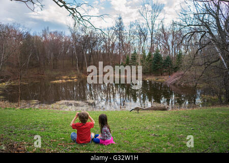 Boy and girl sitting by lake looking at goose with binoculars Stock Photo