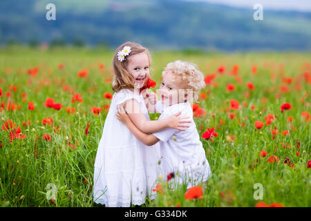 Little curly blond boy and girl play in poppy flower field. Child picking red poppies. Toddler kid in summer meadow. Stock Photo