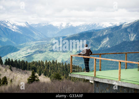 Young man in hood looking on the mountain landscape from wooden pier at morning. Rear view photo. Stock Photo