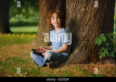 The boy of 8-9 years sits in park under a tree with the tablet in hands. The fellow with a blond curly hair looks in a camera Stock Photo