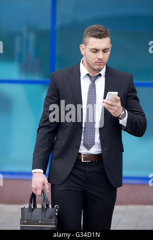Portrait of serious young business man looking at smartphone screen while walking outdoors. Handsome caucasian man using mobile Stock Photo