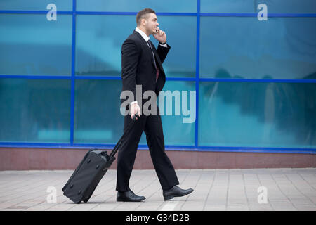 Profile view portrait of cheerful young man on business trip walking with his luggage while talking on smartphone in front Stock Photo