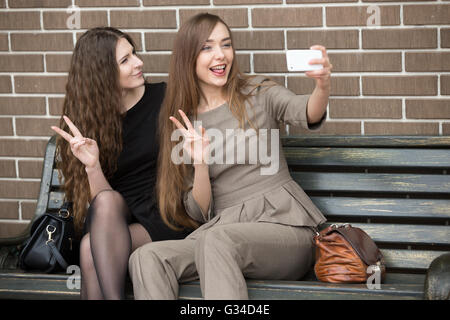 Portrait of two beautiful young girlfriends sitting on bench and taking a selfie on the street. 2 Happy attractive women friends Stock Photo