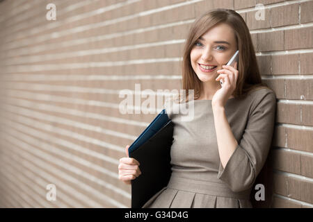 Waist portrait of cheerful young woman standing in the street beside brick wall, talking on smartphone. Happy beautiful Stock Photo
