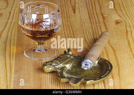 Burning cigar, cognac in a crystal glass and old brass ashtray on a wooden table. Stock Photo