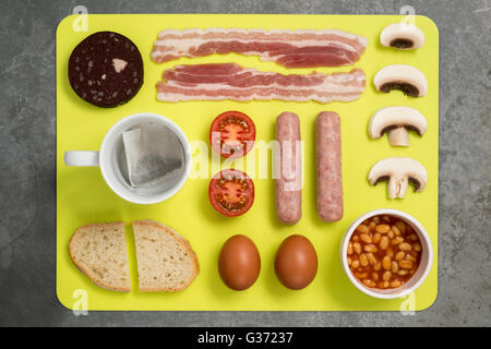 Tea bag in cup and a selection of cooked English breakfast laid flat on a yellow dining mat laid on a grey slate surface. Stock Photo