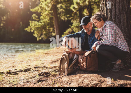 Mature couple stops to check their position by using a compass while out on hike in the forest. Man and woman sitting by a tree Stock Photo