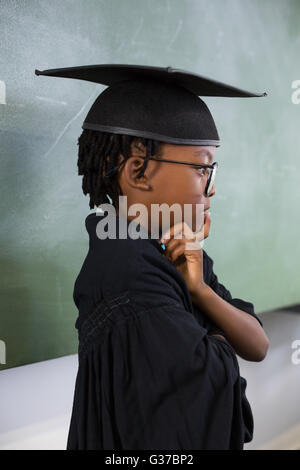 Thoughtful schoolboy wearing graduation gown in classroom Stock Photo