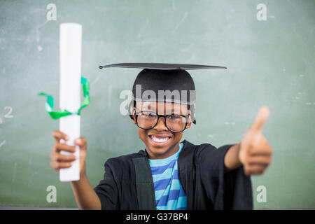 Schoolboy in mortar board holding certificate and showing thumbs up in classroom Stock Photo