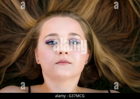 A beautiful teenage girl lays on the ground with her long, blond hair spread out around her.  She looks blankly up at the camera Stock Photo