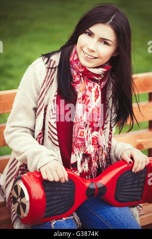 Female model holding modern red electric mini segway or hover board scooter in hands while sitting on a bench in green park.Girl wearing trending boho style clothes. Stock Photo